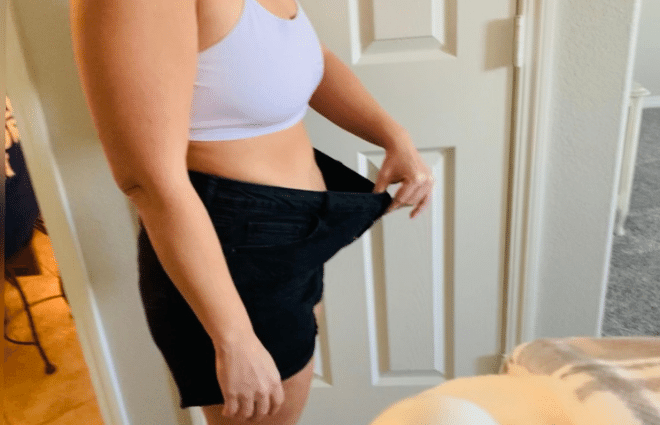Chrystal in sports bra and old, loose workout shorts, holding waistband of shorts out to show how much weight and girth she has lost.